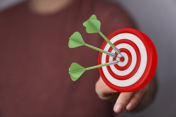 Close-up of a hand holding a dartboard with three darts hitting the center, conveying the ideas of...