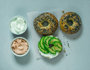Two everything bagels isolated on a flat lay background, one prepared with cream cheese and sliced avocado. Breakfast background.