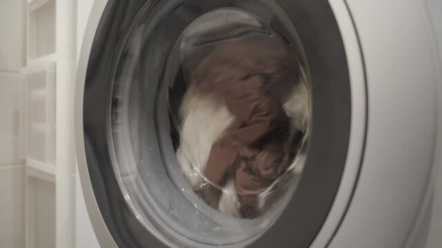 Bed linen tumbles inside the metal-and-glass drum of the washing machine. The low-speed rotation of the appliance creates a repetitive thumping and wet sloshing sound.