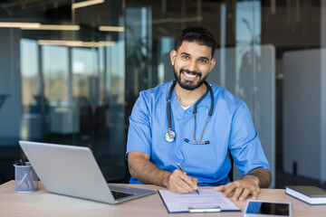 Obraz premium Young male doctor or medical student in blue scrubs and stethoscope smiling at the camera while writing notes on paper, sitting at a desk with a laptop in a modern office