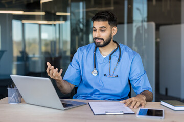 Male doctor in blue scrubs and stethoscope providing a telehealth video call consultation, offering healthcare advice from his office with a laptop and documents on the desk