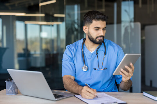 Male healthcare professional in blue scrubs and stethoscope reviewing patient information on a digital tablet while simultaneously writing notes on a clipboard, at an office desk with a laptop