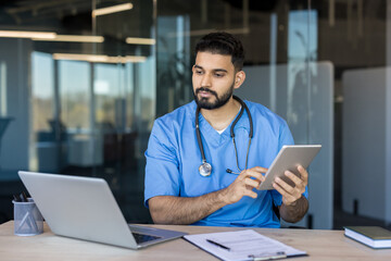 Young male doctor wearing blue scrubs and stethoscope, sitting at a desk and actively using a digital tablet for healthcare data in a clean, contemporary office environment