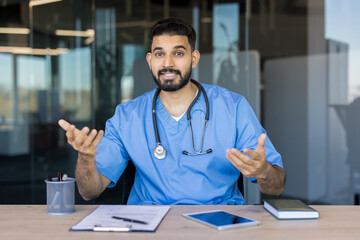 Male doctor in blue scrubs and stethoscope working at a desk, looking at the camera and gesturing while providing telemedicine service or healthcare advice to patients
