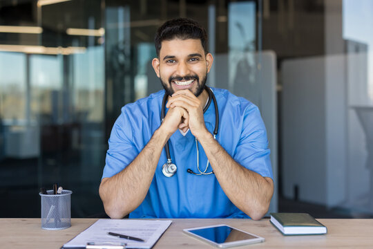 Young indian male doctor wearing scrubs and a stethoscope, sitting at a desk and smiling confidently, embodying professional medical care and support in a clinic environment