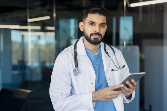 Male indian doctor wearing a white lab coat and stethoscope, confidently holding a digital tablet in a bright, contemporary medical facility, representing innovation and professional healthcare