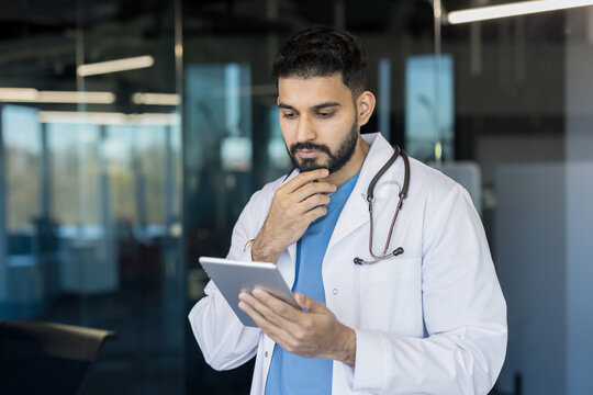Young male doctor in white lab coat and stethoscope thinking. Reviewing medical information on a digital tablet in a modern clinic or office setting. Representing innovation and professional analysis