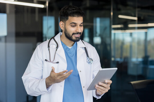 Indian male doctor in white coat with stethoscope offering virtual telemedicine consultation via tablet, smiling and gesturing while providing remote healthcare advice and support