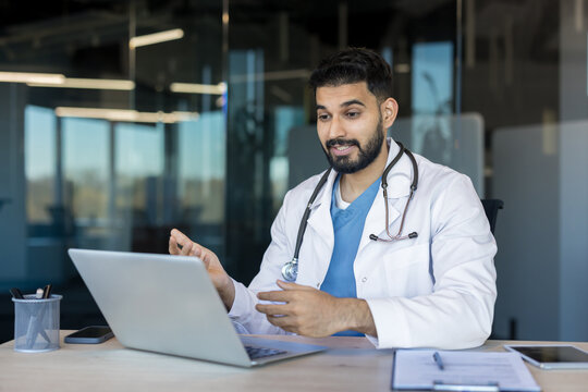 Indian male doctor in white lab coat and stethoscope consulting patient during virtual medical appointment, sitting at desk in modern office, offering healthcare advice online