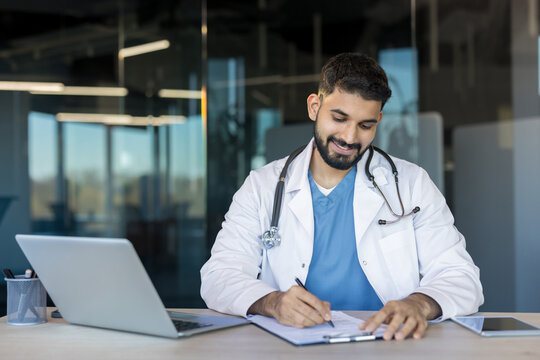 Young male doctor in a white coat with stethoscope smiling while writing patient notes on a clipboard at a modern clinic desk with laptop and tablet nearby, symbolizing care