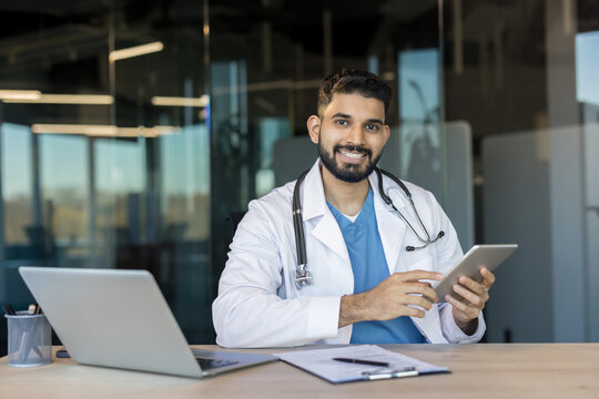 Indian male doctor wearing medical uniform smiling looking at camera, sitting at desk with laptop and documents, holding a digital tablet for healthcare technology and patient care
