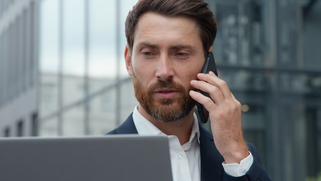 Serious Caucasian mature business man talking phone looking at laptop in city middle-aged businessman male manager consultant talk smartphone call consulting client checks information in computer