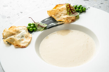 A bowl of creamy, elegant soup in a white dish, garnished with green peas, crispy crackers, and...