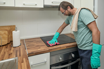 Man cleaning electric stove in modern kitchen with gloves and sponge. Person washing and wiping cooktop with detergent and cloth, doing household chores and keeping kitchen clean. Concept of home