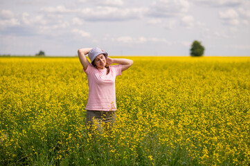 Woman in a bucket hat stands in yellow rapeseed field, enjoying the warmth and peace of a sunny summer day