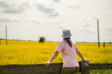 Young woman wearing a bucket hat leaning on an old wooden fence enjoying a warm summer day against a rustic farmstead surrounded by blooming rapeseed fields
