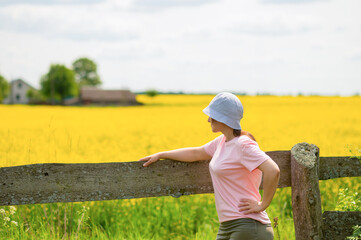 Young woman wearing a bucket hat standing by an old wooden fence looking into the distance at a rustic farmstead in a blooming rapeseed field