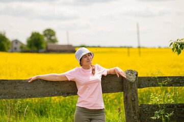 Young woman wearing a bucket hat leaning on an old wooden fence enjoying a warm summer day against a rustic farmstead surrounded by blooming rapeseed fields
