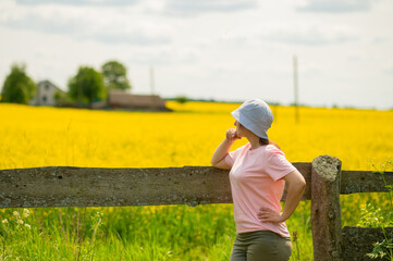 Young woman wearing a bucket hat leaning on an old wooden fence enjoying a warm summer day against a rustic farmstead surrounded by blooming rapeseed fields