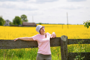Young woman wearing a bucket hat leaning on an old wooden fence enjoying a warm summer day against a rustic farmstead surrounded by blooming rapeseed fields