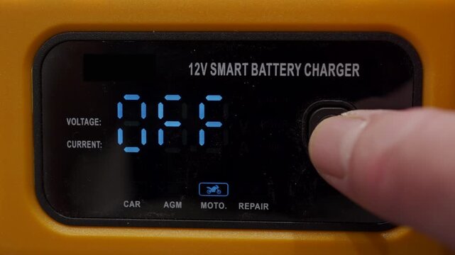 A finger presses a button on the plastic car battery charger control panel to change the charging mode. This action is accompanied by a short, soft clicking sound.