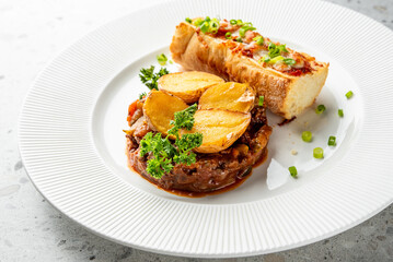 Hearty meat and vegetable stew with golden roasted potatoes, topped with fresh herbs, and served with a cheesy baguette slice on a white plate