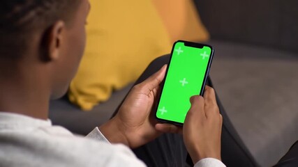 Over the shoulder shot of an African American man using a smartphone with a green screen for easy keying browsing content or social media at home. - Powered by Adobe