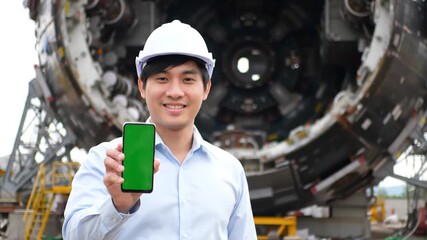 Smiling Asian engineer in a hard hat proudly displays a smartphone with a green screen standing in front of heavy industrial machinery at a construction site showcasing modern technology in engineeri. - Powered by Adobe