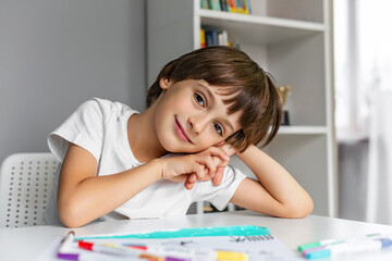 Smiling child resting at a desk with drawing supplies in a creative study space.