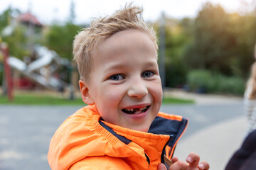 Portrait happy smiling boy with missing tooth wearing bright orange jacket at playground. Joyful expression and sunlight in background create cheerful childhood mood full of energy and spontaneity