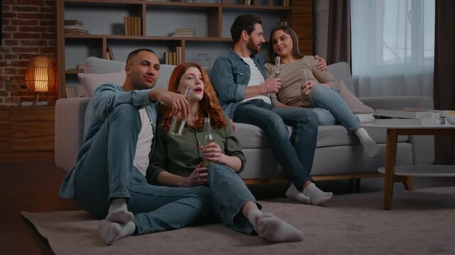 Two diverse married couple four multiracial friends watching television at home party drinking beer alcohol cider drink in living room multiethnic multiracial men women focused enjoying movie film tv