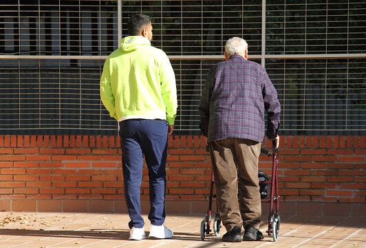 Anciano con andador ortop&eacute;dico y joven ayudante y cuidador de persona mayor caminando de espaldas en patio