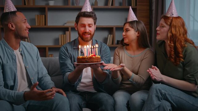 Four multiracial friends at birthday party singing congretulate man with cake trying blowing candles Latina American guy stopping saying no laughing together women men happy celebration friendship
