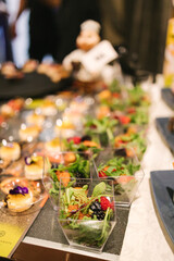 Elegant buffet setup with fresh salad mixes in transparent square cups, featuring greens, berries, and crispy toppings. Accompanied by delicate desserts with edible flower garnish. Perfect for a sophi