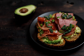 Close-up of avocado toast with salami, prosciutto, and green onions on a dark plate, healthy food and a satisfying meal.