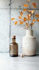 A still life composition featuring autumn foliage in a vase, a glass bottle, and a nut, set against a textured concrete background.