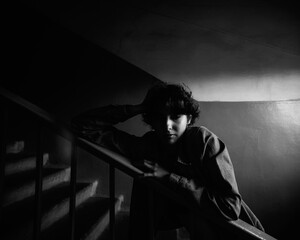 Young woman with tattoos sitting on stairs in a dimly lit corridor, showcasing urban lifestyle and introspective mood in a monochrome setting