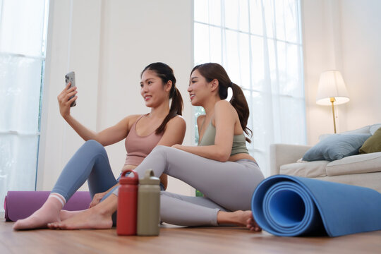 Two Asian women in yoga outfit smiling while taking selfie together after exercise, showing friendship and healthy lifestyle at home.