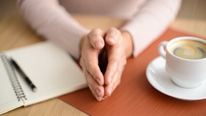 Woman's hands resting on a table, praying or contemplating, reflecting on life issues, finding solace and peace, surrounded by a notebook, pen, and a cup of coffee