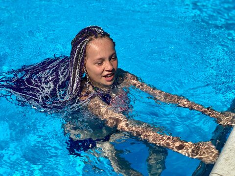 Teen girl with purple and white afro braids swims near the pool edge, holding the rail while sunlight reflects across the clear blue water. Her wet braids float behind her, creating a bright, joyful - Powered by Adobe