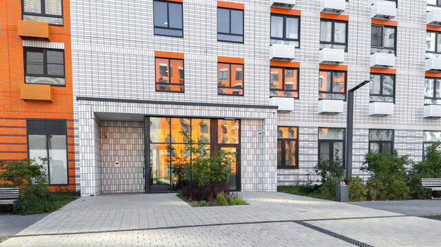 Entrance of modern residential building with geometric tiled facade in white and orange tones, large reflective windows, landscaped courtyard and urban design concept for real estate housing projects - Powered by Adobe