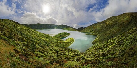 A clear lake surrounded by green hills and mountains under a blue sky with clouds on a sunny day, crater lake Lagoa do Fogo, Caldeiras, Sao Miguel Island, Azores, Portugal