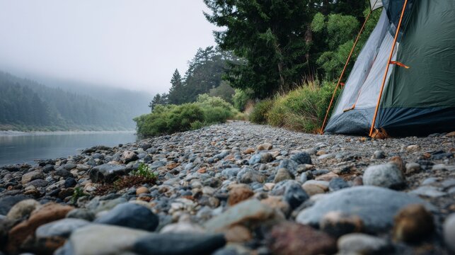 Tent pitched on a rocky beach near water with distant forest in misty weather 85 characters