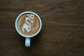 A person holding a cup of coffee with latte art showing the number "2026," wearing a pink sweater and relaxing indoors.