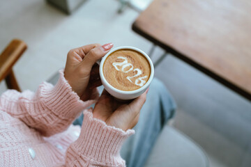A person holding a cup of coffee with latte art showing the number "2026," wearing a pink sweater and relaxing indoors.