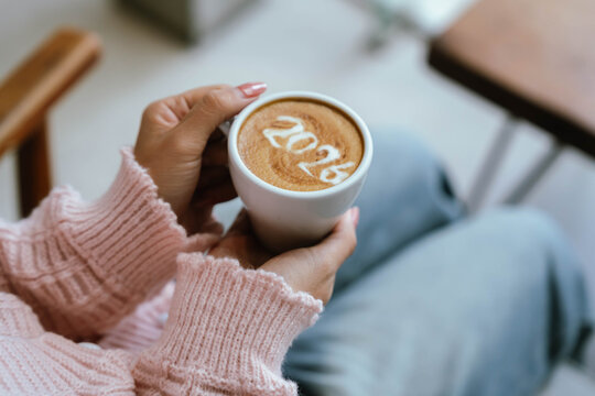 A person holding a cup of coffee with latte art showing the number "2026," wearing a pink sweater and relaxing indoors.