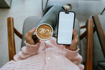 A person holding a cup of coffee with latte art showing the number "2026," wearing a pink sweater and relaxing indoors.