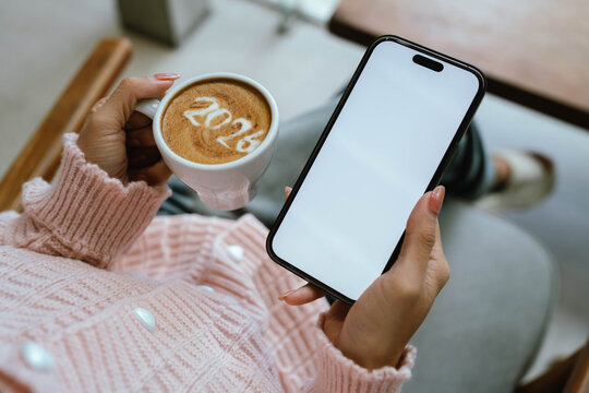 A person in a cozy pink sweater holding a cup of coffee with "2026" latte art while using a smartphone with a blank screen, symbolizing a fresh start for the new year.