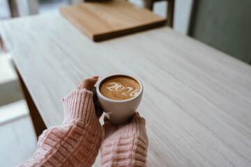 A person holding a cup of coffee with latte art showing the number "2026," wearing a pink sweater and relaxing indoors.