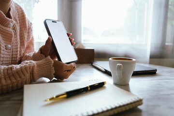 Woman using smartphone at cozy workspace with laptop, notebook, and coffee cup on marble table in soft daylight atmosphere.
